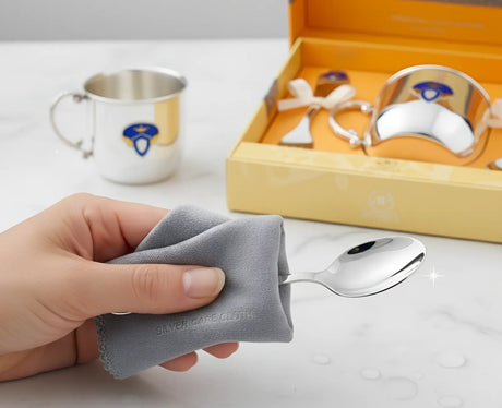 Hand poliert einen silbernen Babylöffel mit einem speziellen Silberpflegetuch. Im Hintergrund ein Baby-Geschenkset aus Silber.