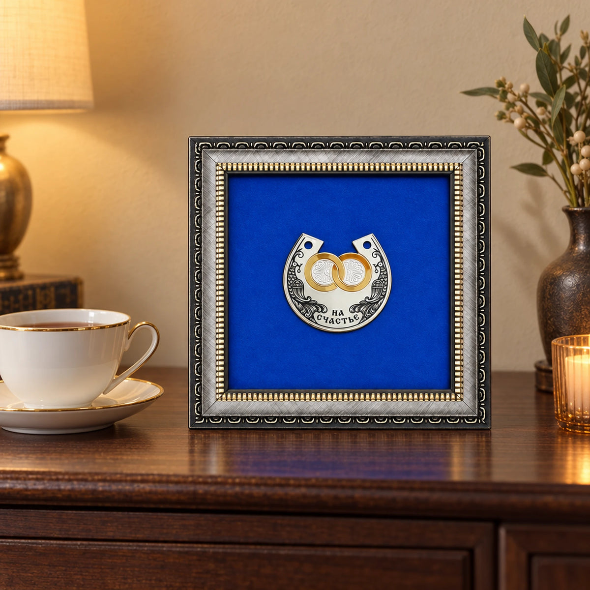 Wandbild Hufeisen mit vergoldeten Eheringen 925 Silber, 15 × 15 cm, auf einer dunklen Kommode neben einer Teetasse