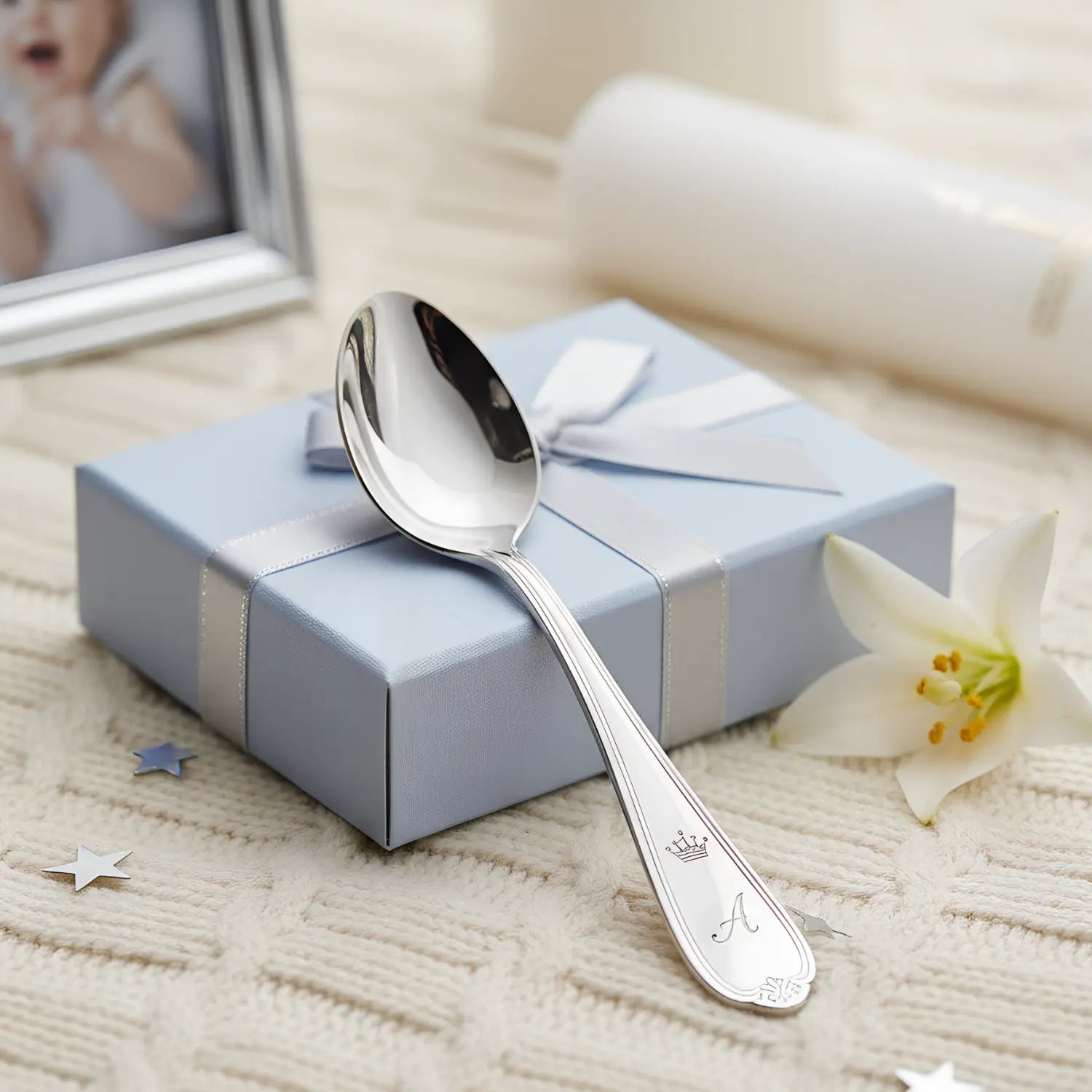 Eleganter Babylöffel aus 925 Sterling Silber mit Gravur, präsentiert auf einem weichen Stricktuch neben einer blauen Geschenkbox und einem Babyfoto.