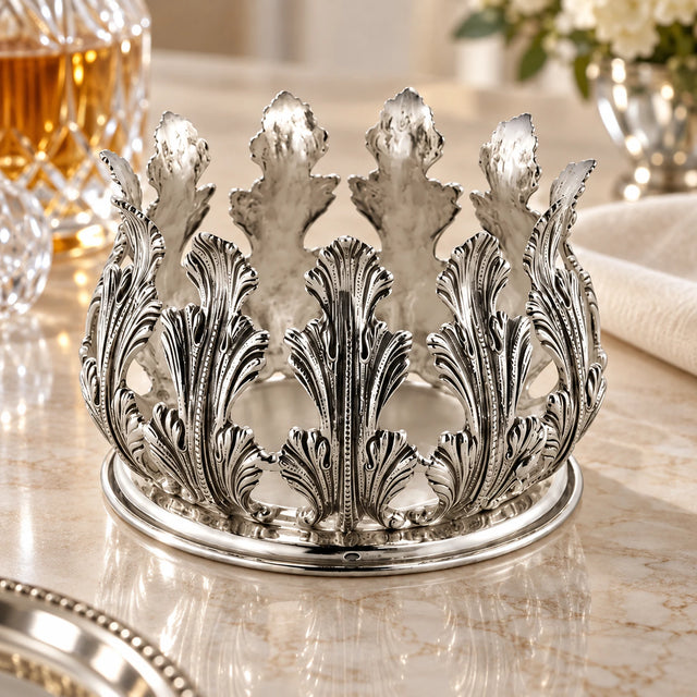 Flaschenuntersetzer aus 925 Silber mit Kranz aus getriebenen Akanthusblättern auf heller Marmorplatte, daneben Kristallglas und Leinenserviette — Lifestyle-Aufnahme