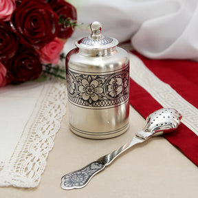 Silberne Teedose mit floralem Niello-Dekor und passendem Teesieblöffel, stilvoll dekoriert auf edler Tischdecke.