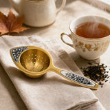 Teesieb Aurora Gold 925 Silber vergoldet auf Leinenserviette neben dampfender Teetasse und losem Tee — Lifestyle